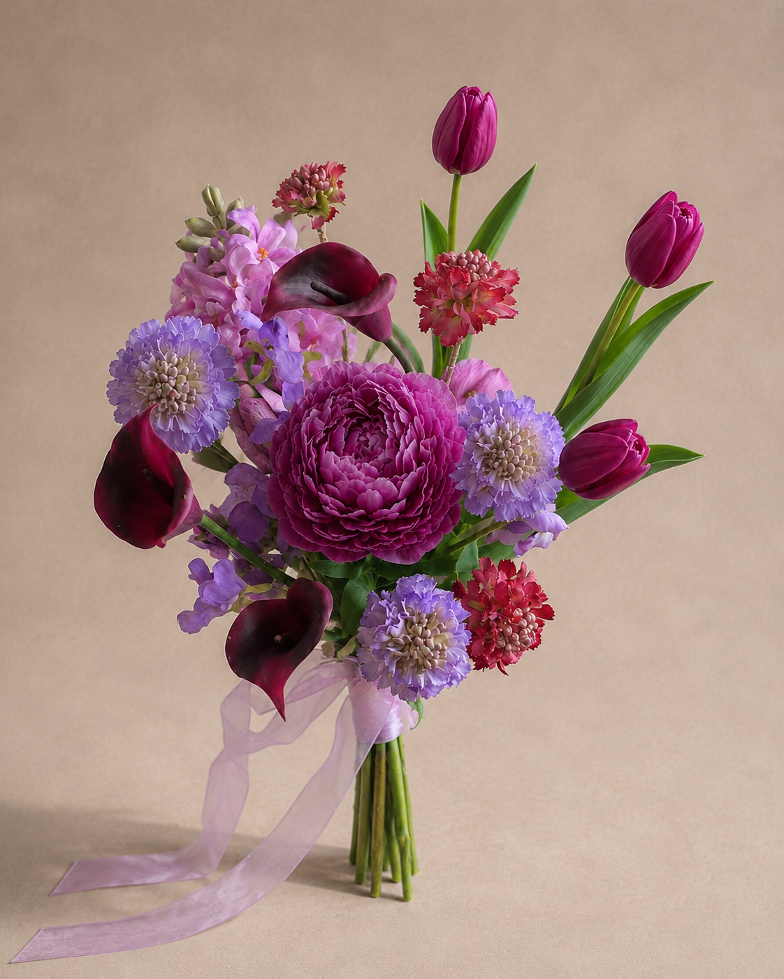 Bride holding Jewel Peony Posy close to her chest indoors, featuring burgundy calla lilies, purple scabiosa and jewel-tone florals