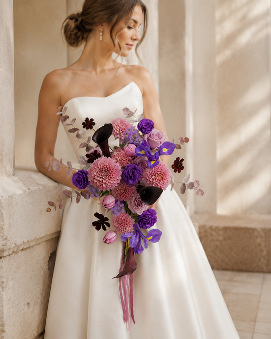 Bride in an ivory wedding gown holding the Twilight Dahlia Cascade bouquet with dusty pink dahlias, violet irises and black calla lilies in a warm stone interior