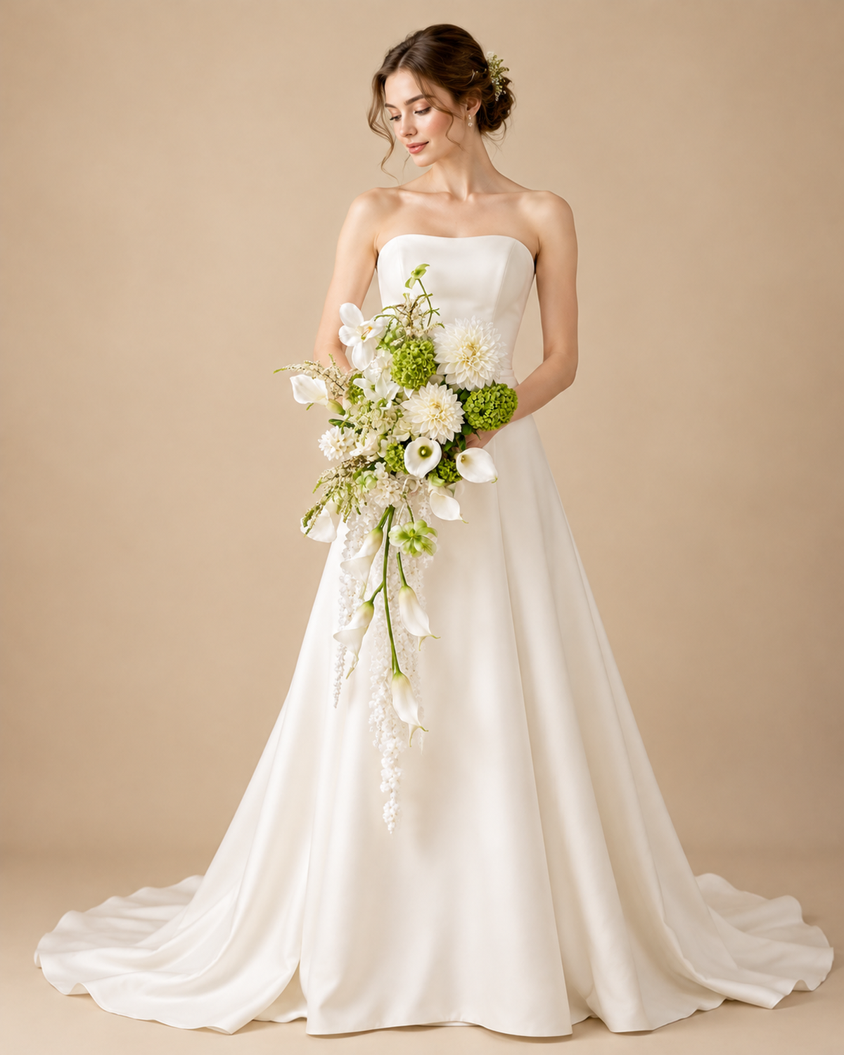 Bride in a strapless ivory gown holding the White Green Cascade bridal bouquet of white dahlias, white calla lilies and green hydrangea with trailing white blooms, against a beige studio background; the bride and gown are shown for display and are not included.