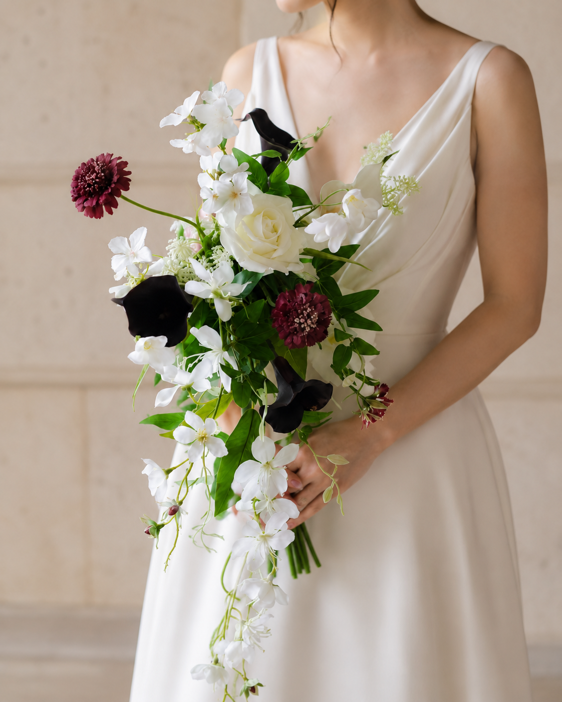 Bride in a spaghetti-strap ivory wedding gown standing among stone columns and cypress trees, holding the Ivory Noir bridal bouquet